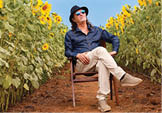 A person wearing a dark hat, sunglasses, a dark blue shirt, and light-colored pants is sitting on a wooden chair with legs crossed in the middle of a sunflower field under a clear sky. Description generated by AI