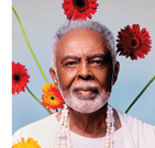 A person with short curly hair wearing layered white beaded necklaces and a light-colored top is standing in front of a light blue background decorated with vibrant red and orange flowers. Description generated by AI