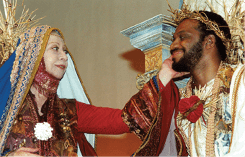 Two adults dressed in ornate, historical or theatrical costumes are smiling at each other, with one gently touching the other's face. Description generated by AI