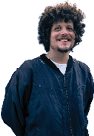 A person with curly hair is smiling and wearing a dark zip-up jacket over a white shirt while standing against a light blue wall. Description generated by AI