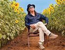 A person wearing a dark hat, sunglasses, a dark blue shirt, and light-colored pants is sitting on a wooden chair with legs crossed in the middle of a sunflower field under a clear sky. Description generated by AI
